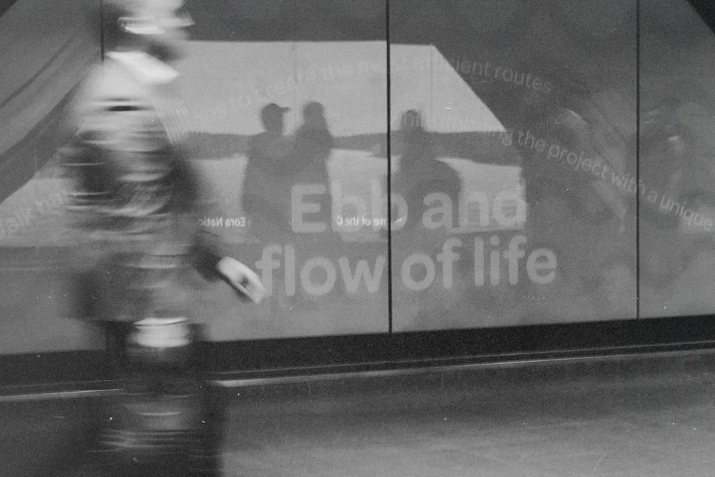 A blurred figure walks past a reflective wall displaying the words “Ebb and flow of life,” with silhouettes visible in the reflection