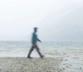 Person walking along a rocky lakeshore on a foggy day with mountains fading into the mist
