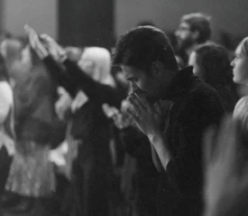Group of people standing closely together in a dimly lit room with several individuals raising their hands and one person in the foreground holding their hands together in front of their face