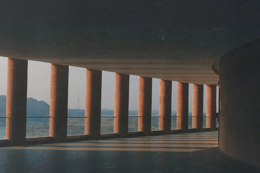 Long covered walkway with tall red columns casting angled shadows across the floor, overlooking a distant landscape in soft morning light