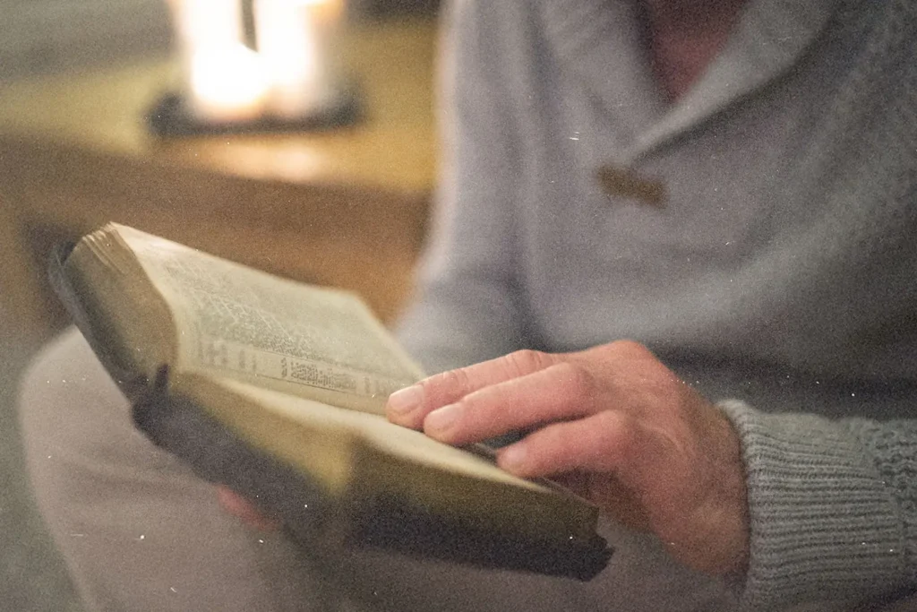 Person holding an open Bible in soft, warm indoor lighting, reading from its pages