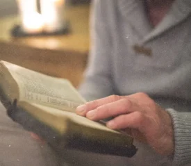 Person holding an open Bible in soft, warm indoor lighting, reading from its pages