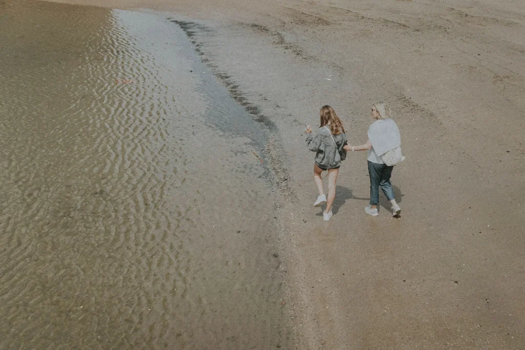 Two people walk side by side along the wet sand near the water’s edge, their reflections visible in the shallow ripples
