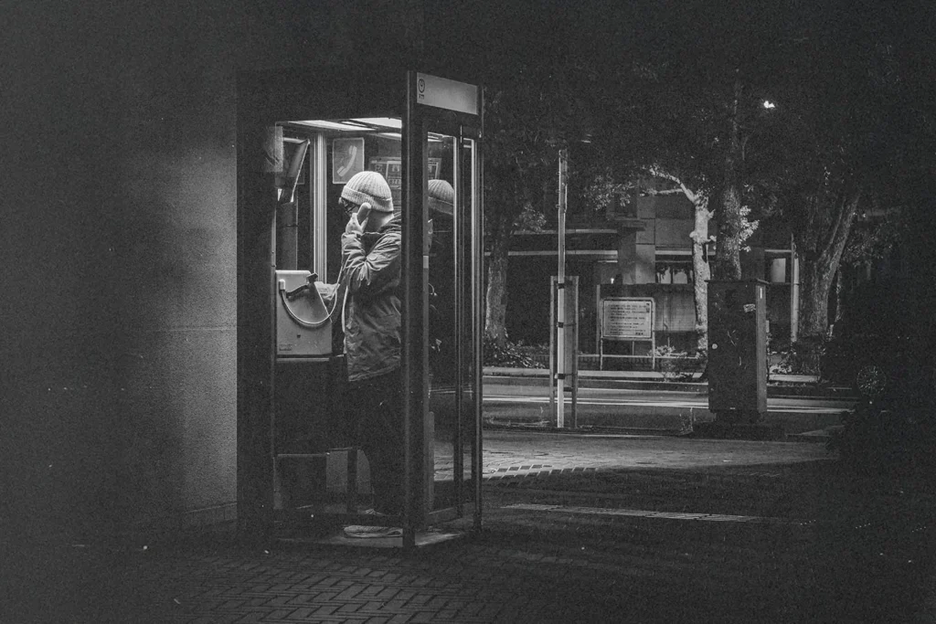 Person standing in a lit phone booth at night on a quiet street