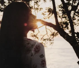 Silhouetted person standing by trees at sunrise, eyes closed as warm light shines through branches, creating a peaceful, reflective moment by the water.