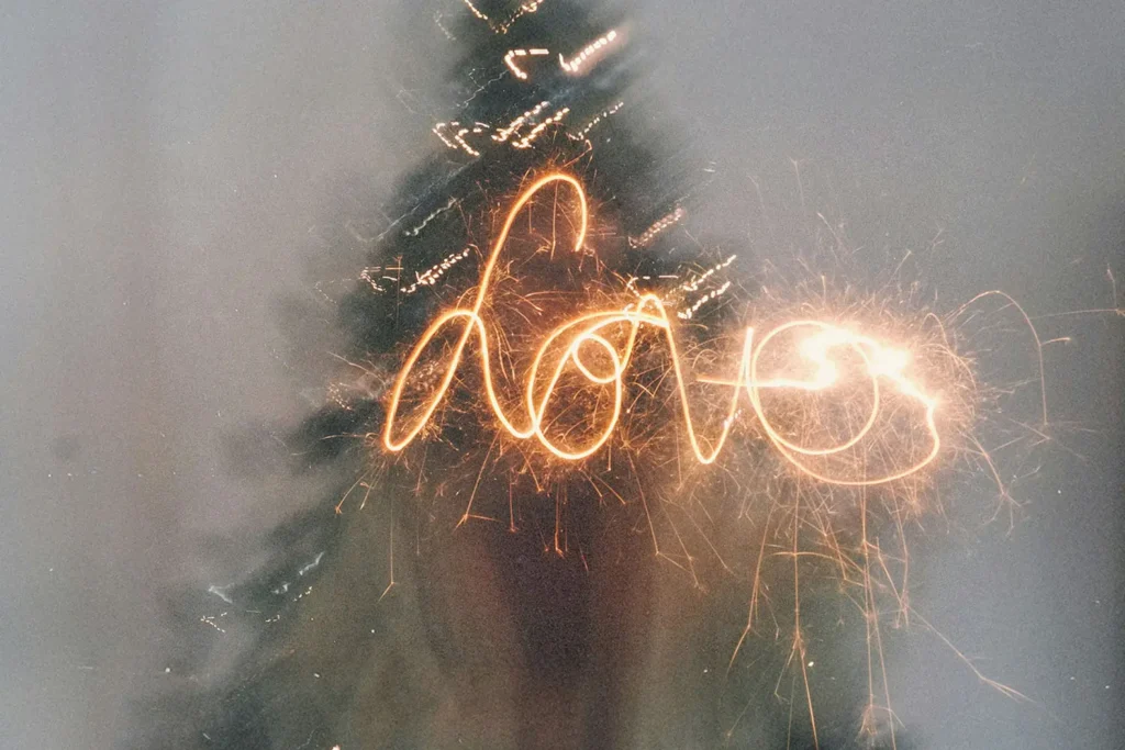 A sparkler writes the word “love” in glowing light trails in front of a blurred Christmas tree