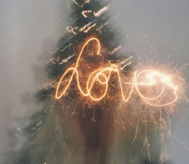 A sparkler writes the word “love” in glowing light trails in front of a blurred Christmas tree