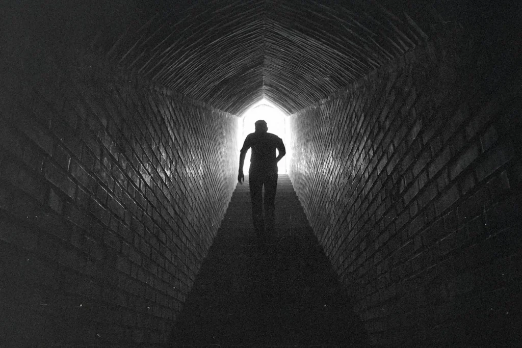 A person walks through a long, dark brick tunnel toward a bright opening at the far end