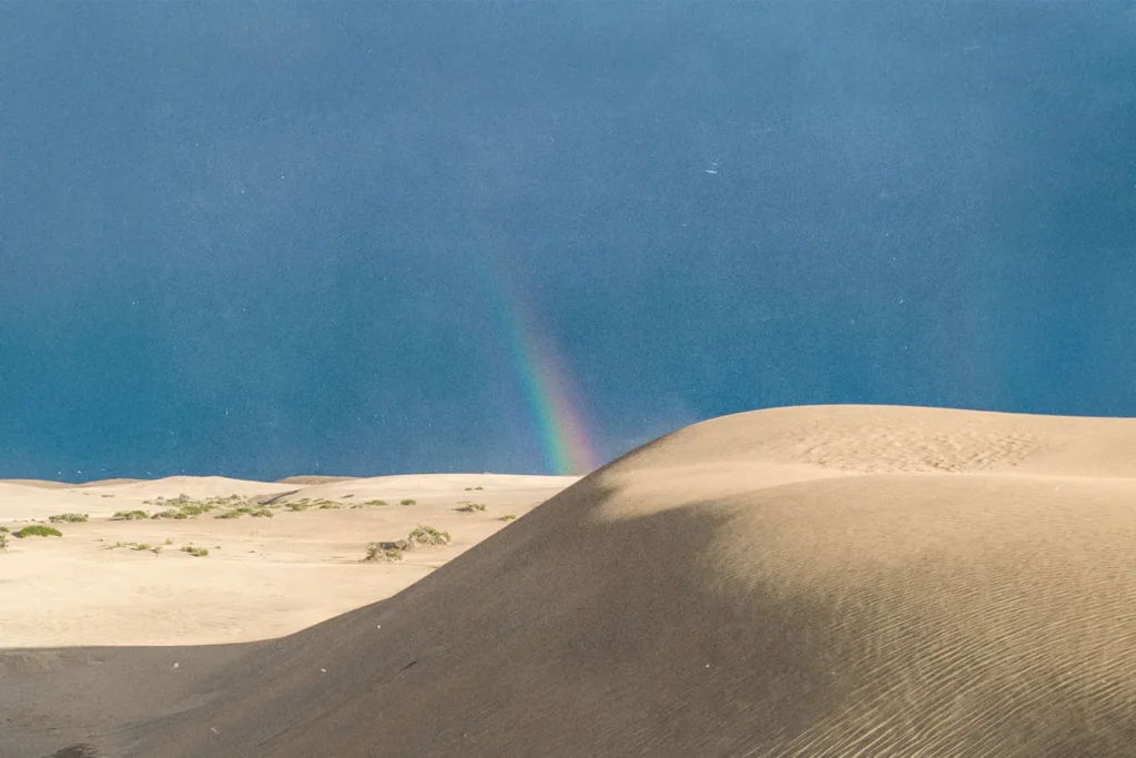 Tall sand dunes under a clear blue sky with a faint rainbow appearing near the horizon