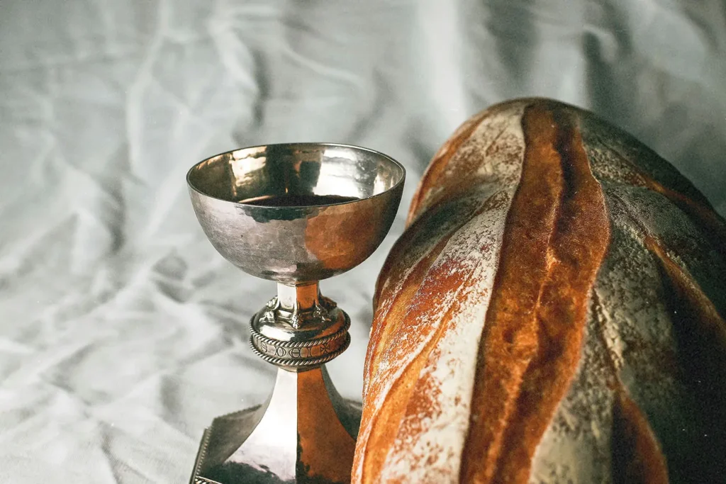 Silver communion cup filled with wine beside a loaf of rustic bread on a soft fabric background