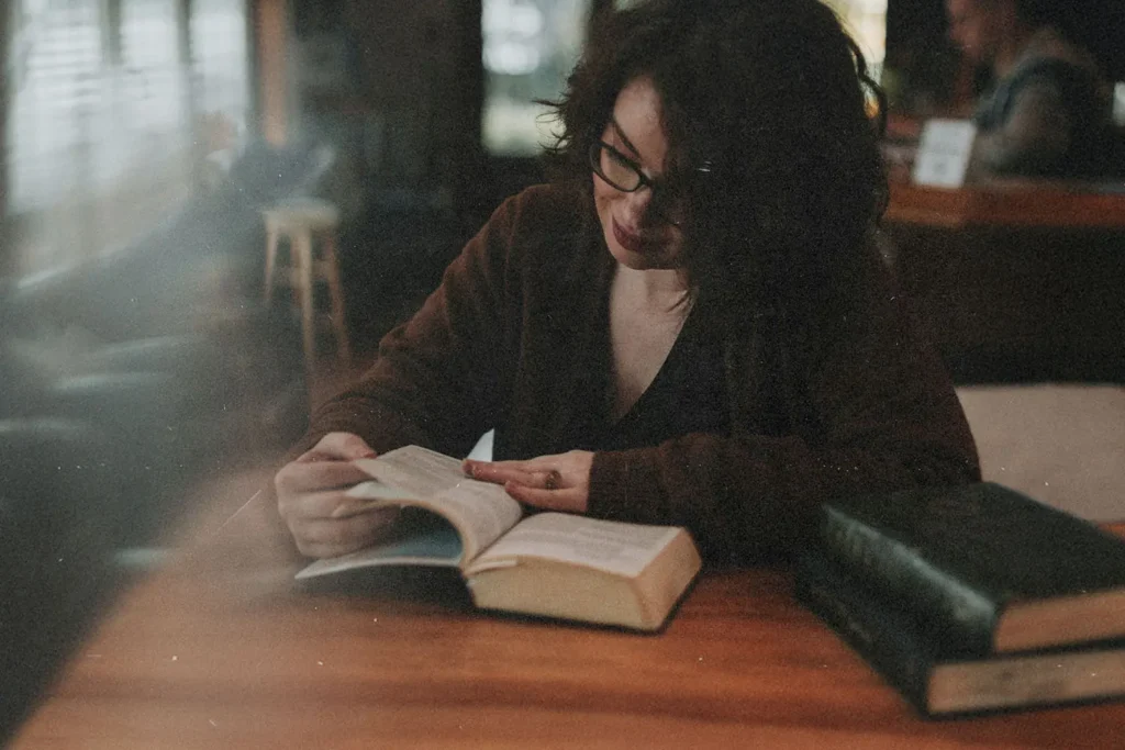 A person sits at a wooden table in a dimly lit room, reading an open book with another large book resting nearby