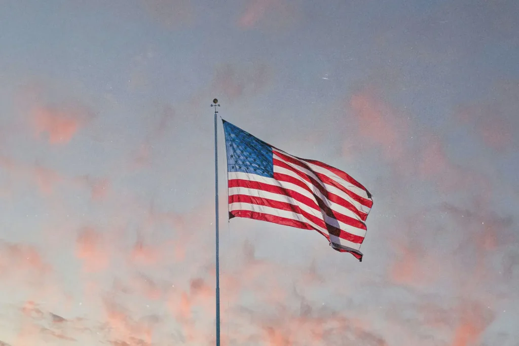 An American flag waves on a tall pole against a pastel evening sky with soft pink clouds
