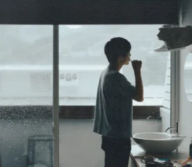 Man brushing their teeth in a small bathroom with a large window overlooking a rainy landscape