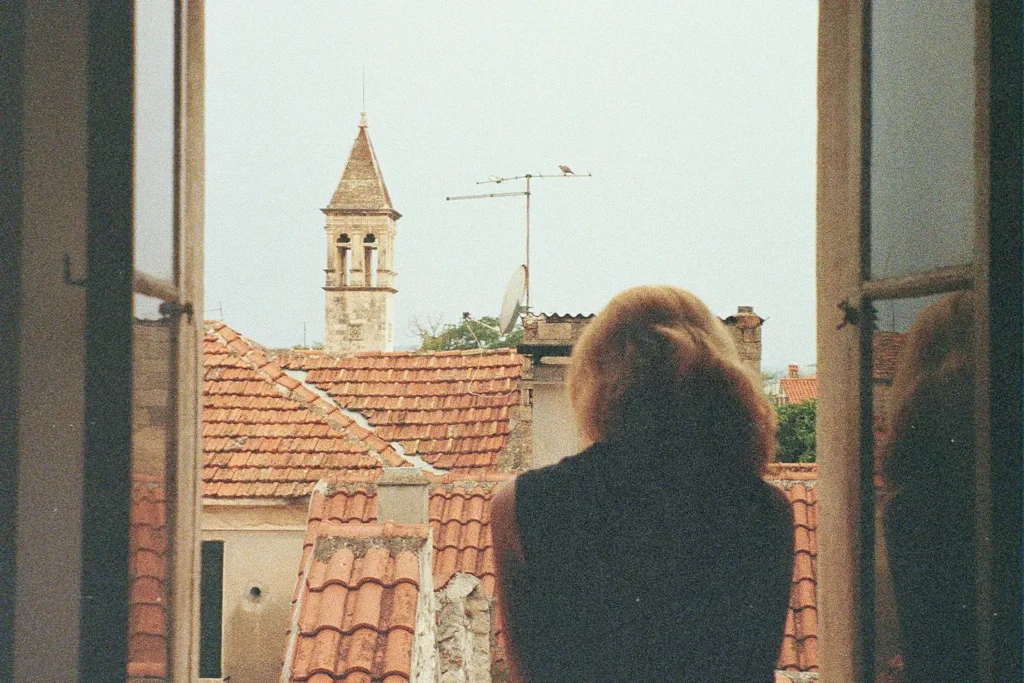 Person stands at an open window looking out over terracotta rooftops and a stone bell tower in a historic town
