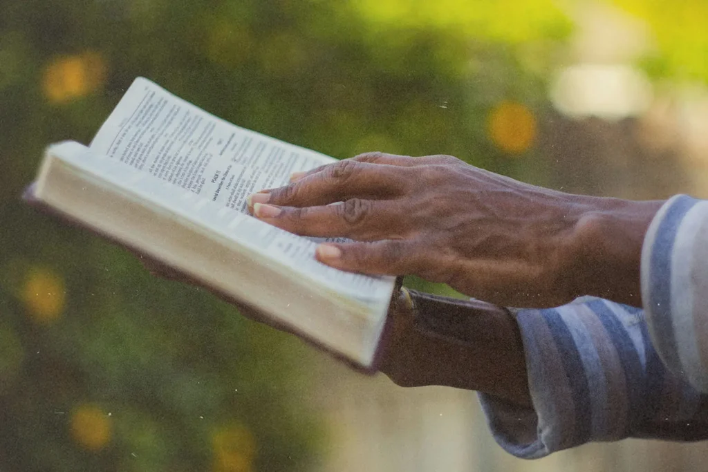 Hand holding an open Bible outdoors, with sunlight illuminating the pages against a soft, green background