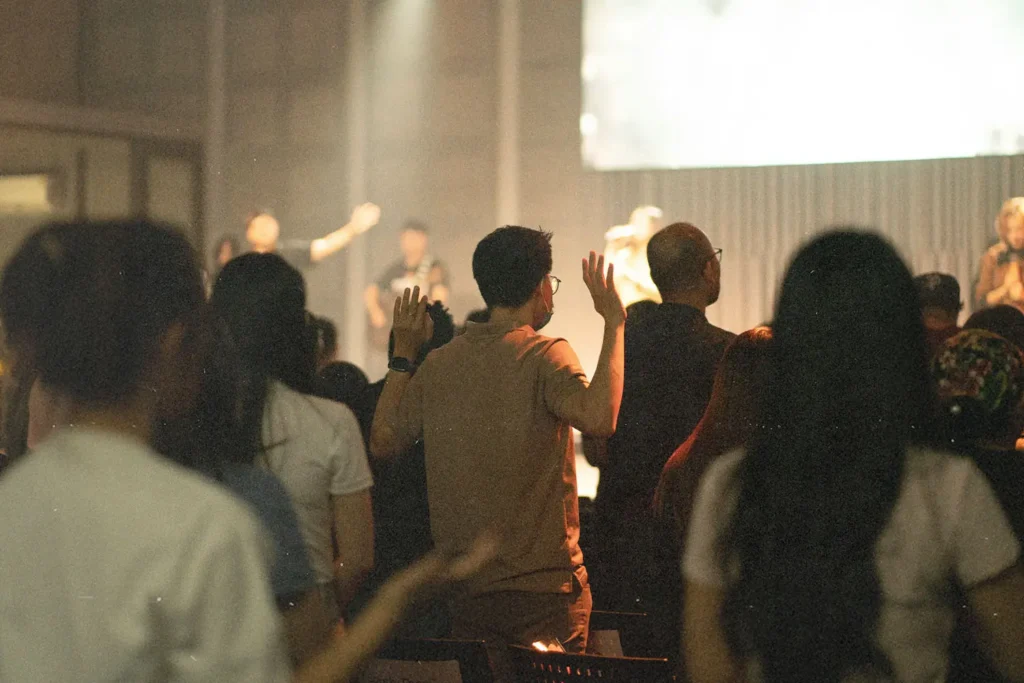 A group of people stand in a dimly lit worship setting with raised hands facing a stage