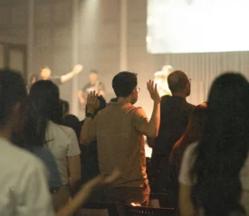 A group of people stand in a dimly lit worship setting with raised hands facing a stage