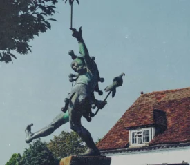 A bronze statue of a lively, dancing figure holding a staff stands in front of a historic building with a red‑tiled roof