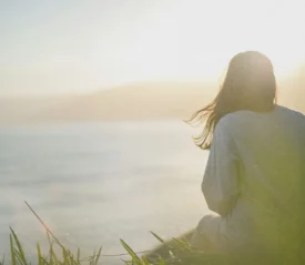 Person sitting in the grass overlooking a calm body of water at sunrise, with sunlight creating a soft, peaceful glow