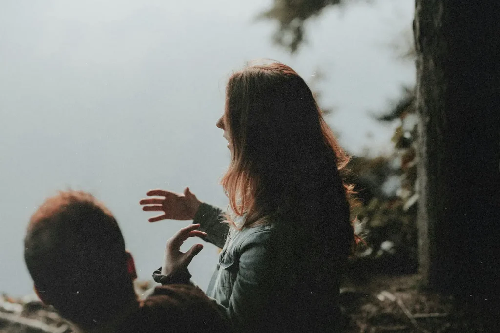 Two people sitting near the edge of a wooded area beside calm water, with one person gesturing while speaking