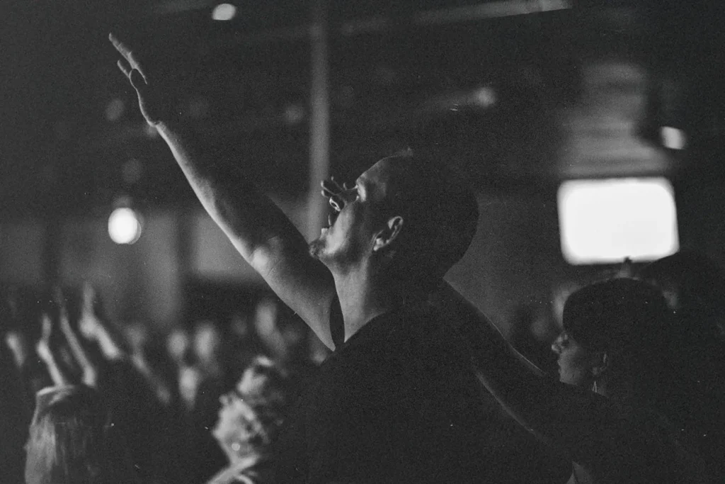 A person in a crowd raises one arm upward in worship or celebration inside a dimly lit room