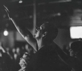 A person in a crowd raises one arm upward in worship or celebration inside a dimly lit room