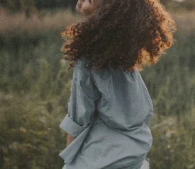Person with curly hair standing in a grassy field with warm sunlight shining from behind