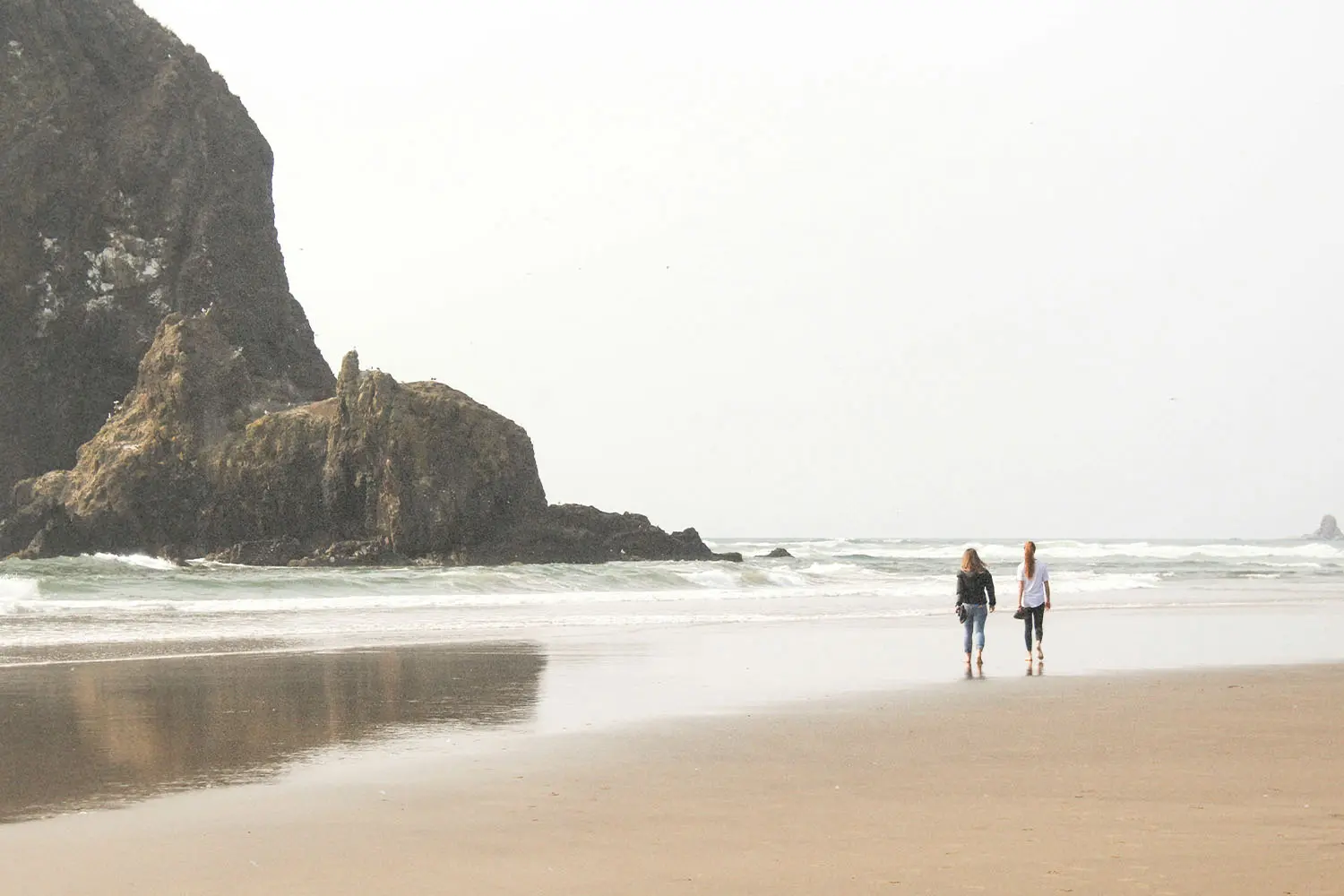 Two people stroll along a wide beach with waves rolling in and a large rocky formation rising from the shoreline nearby