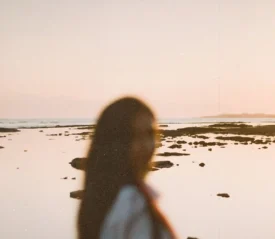A person stands near the shoreline at sunset, with calm water and scattered rocks stretching into the distance
