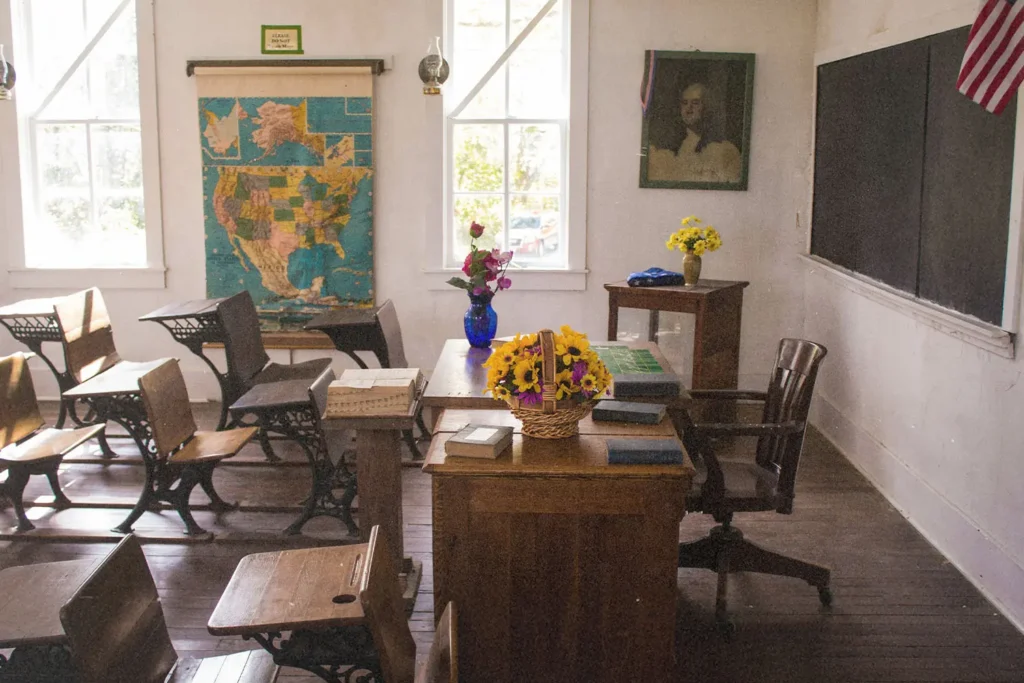 Vintage-style classroom with wooden desks, a teacher’s desk decorated with flowers, a large map on the wall, and sunlight streaming through tall windows