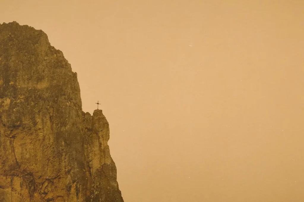 A tall rocky cliff rises against a hazy golden sky, with a small cross standing at the edge of the peak
