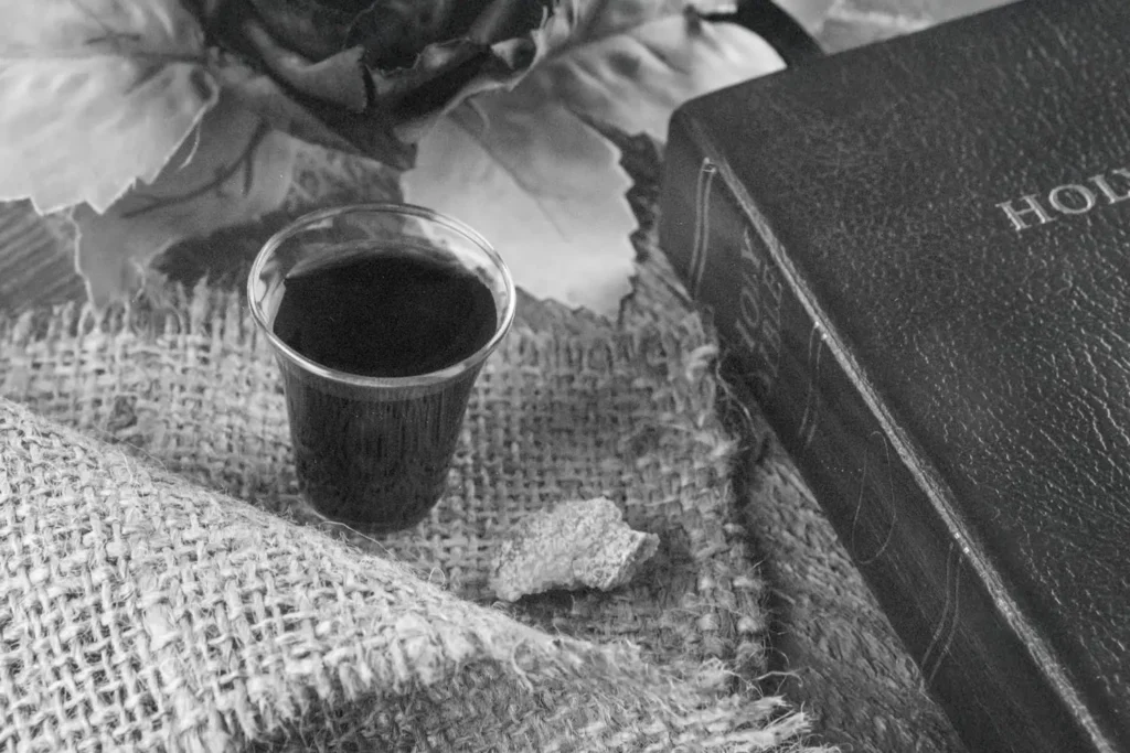 A small cup of communion juice sits on a piece of burlap alongside a broken piece of bread, with a Bible and decorative leaves arranged nearby in a softly lit setting