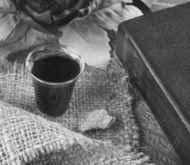 A small cup of communion juice sits on a piece of burlap alongside a broken piece of bread, with a Bible and decorative leaves arranged nearby in a softly lit setting