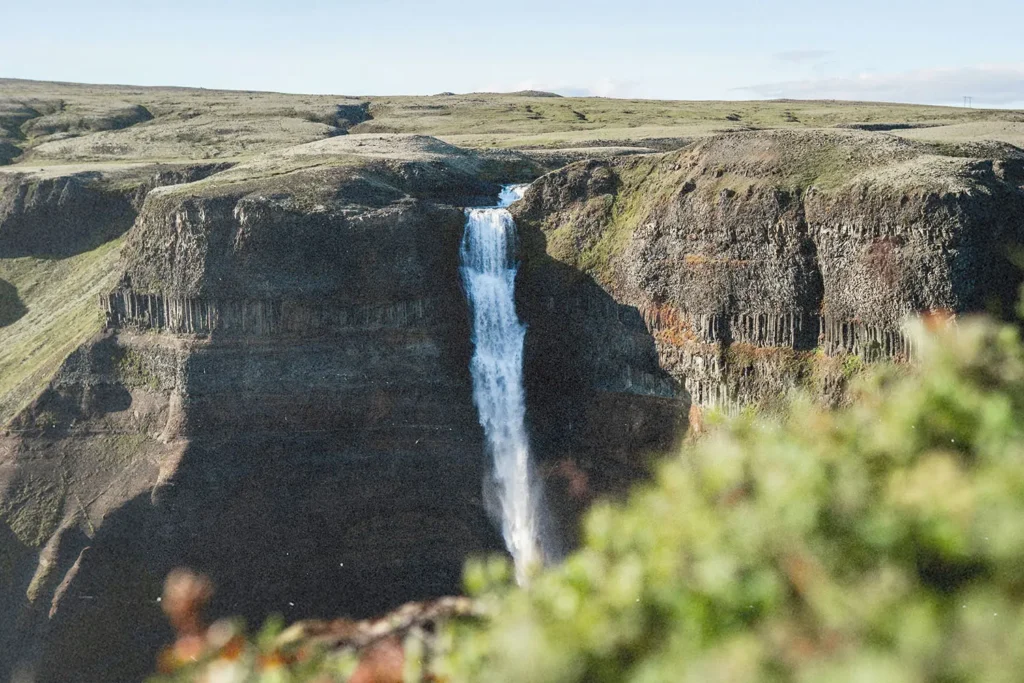 A tall waterfall cascades over a steep cliff into a rocky valley surrounded by green terrain