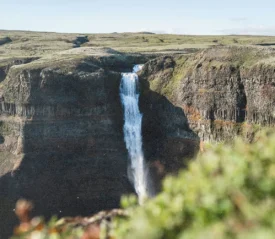 A tall waterfall cascades over a steep cliff into a rocky valley surrounded by green terrain