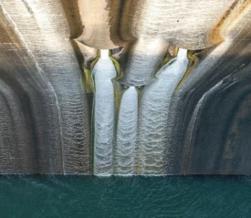 looking down from the top of a dam, water falling down a concrete wall into body of water
