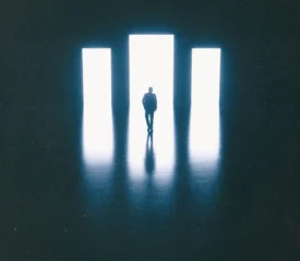Silhouette of a person standing before three tall, bright rectangular doorways in a dark room, with light reflecting across the floor
