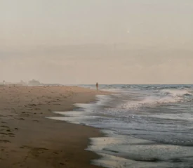 A lone person walks along a quiet shoreline as gentle waves roll onto the sand in the early morning light