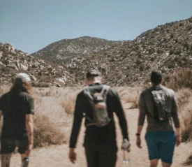 Three people walk along a dirt trail surrounded by dry brush, with rocky mountains rising in the background under a clear sky