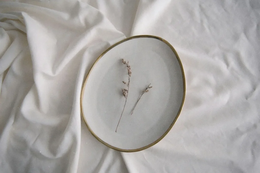 A simple ceramic plate holding two delicate dried sprigs rests on softly wrinkled white fabric