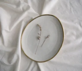 A simple ceramic plate holding two delicate dried sprigs rests on softly wrinkled white fabric