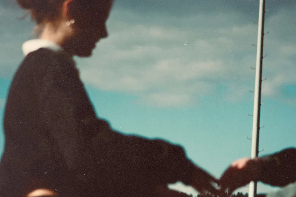 A softly focused outdoor scene showing a person in a dark long‑sleeve top holding hands with another person. A tall flagpole rises into a partly cloudy blue sky in the background.