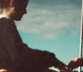A softly focused outdoor scene showing a person in a dark long‑sleeve top holding hands with another person. A tall flagpole rises into a partly cloudy blue sky in the background.
