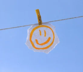 Hand‑drawn smiley face on a torn piece of paper clipped to a clothesline against a clear blue sky