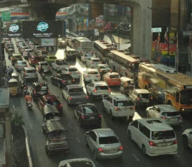Heavy traffic jam on a multi‑lane city street filled with cars, buses, and tuk‑tuks beneath an elevated roadway in a busy urban shopping district