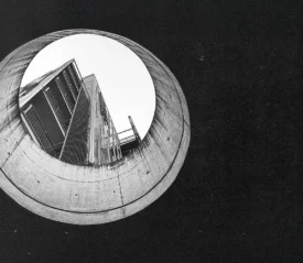 Black‑and‑white photograph looking upward through a large circular concrete opening toward a tall modern building, showing sharp architectural lines and high contrast