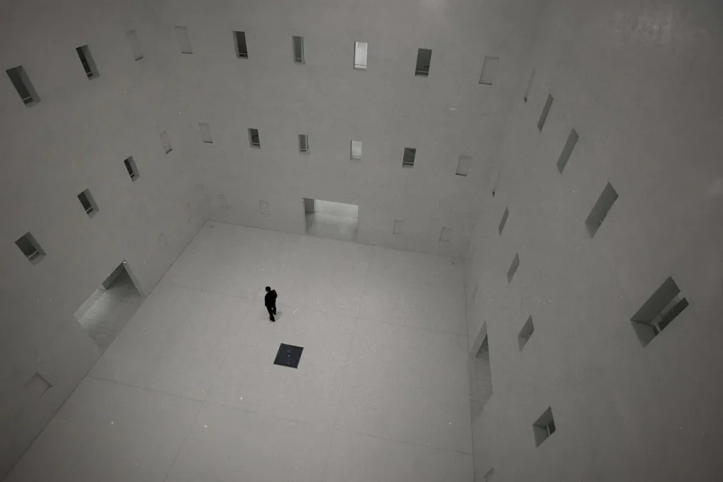 Person standing alone in a large, minimalistic white atrium with tall walls lined with small rectangular openings