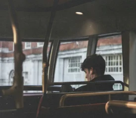 Person seated on the upper level of a bus, looking out large windows at a row of brick and white buildings outside