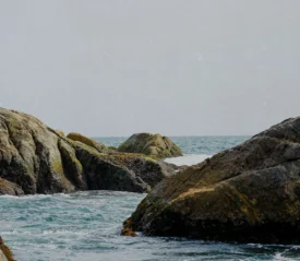 Rocky shoreline with ocean waves moving between large weathered boulders under a pale, hazy sky