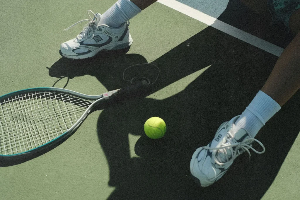 A tennis racket and bright yellow tennis ball rest on a court beside a person wearing athletic shoes and white socks
